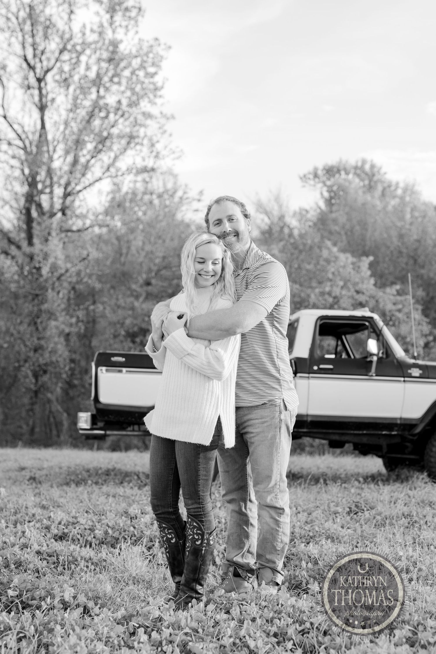A black and white portrait of a couple embracing in front of a classic truck in a grassy field.