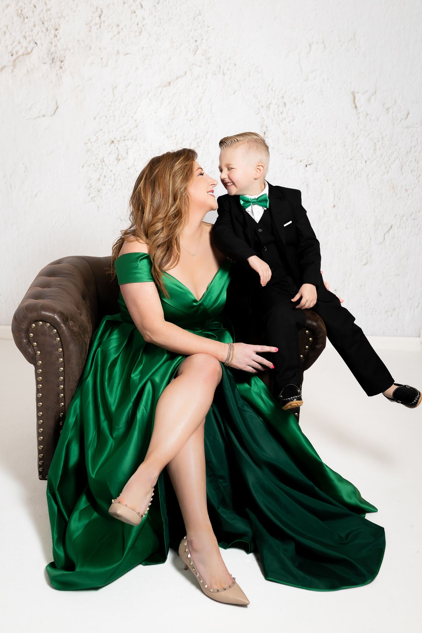 A formal portrait against a white wall features coordinating emerald green and black formal attire on a brown leather sofa.