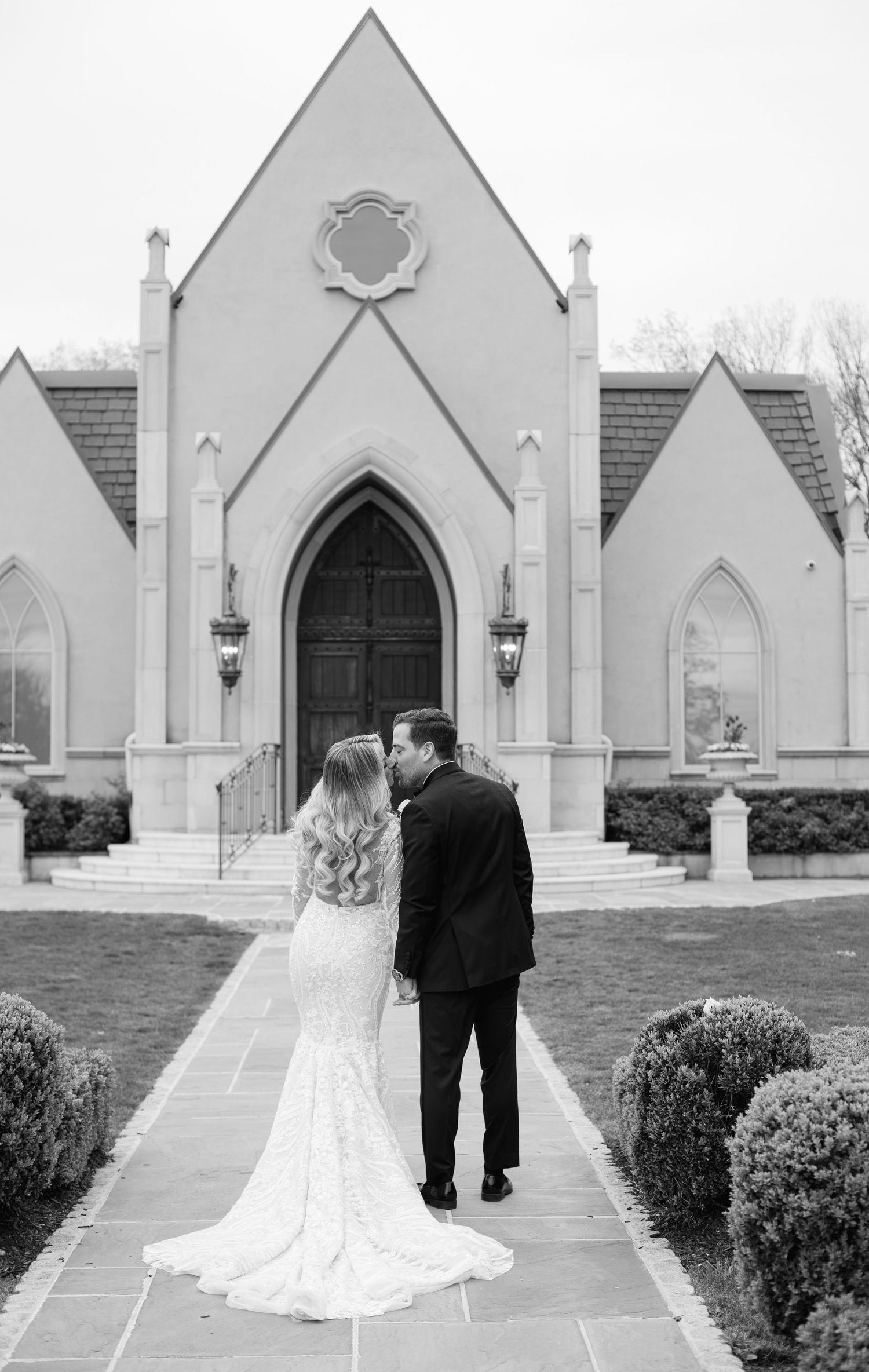 Couple stands before a white gothic revival church in wedding attire.