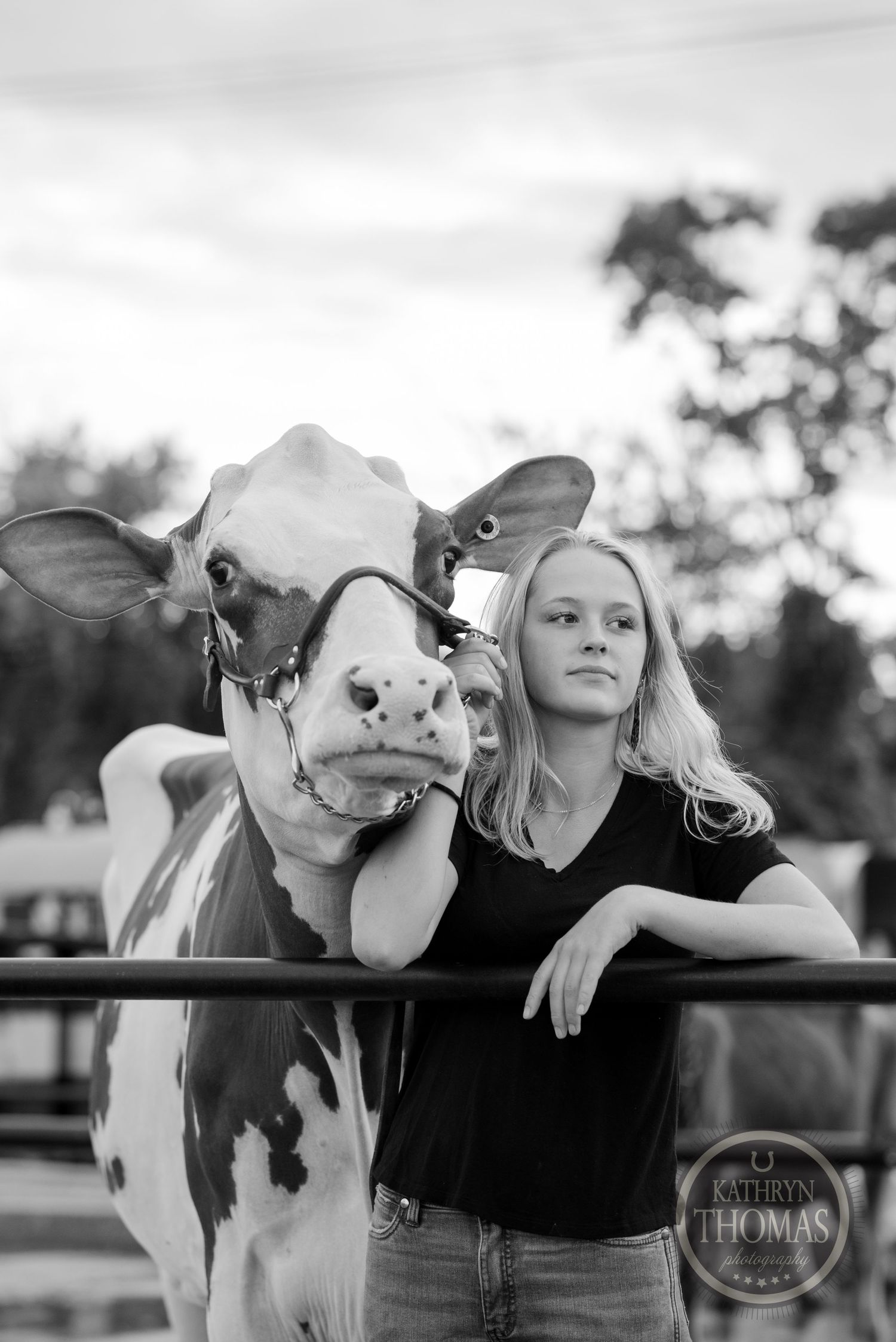 A monochrome portrait shows a dairy cow and handler leaning on a metal fence at dusk.