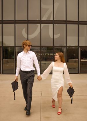 A graduate couple in formal attire holding diplomas while walking outside a university building.