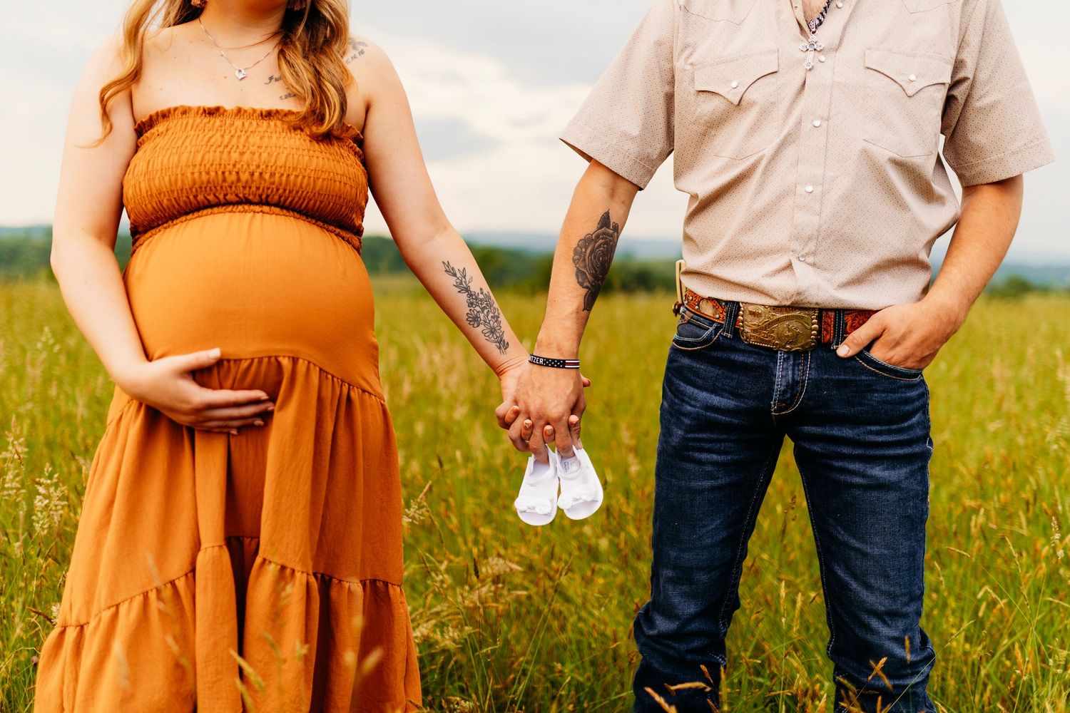 Pregnant couple holding hands in golden field, man holding tiny white baby shoes during maternity session.