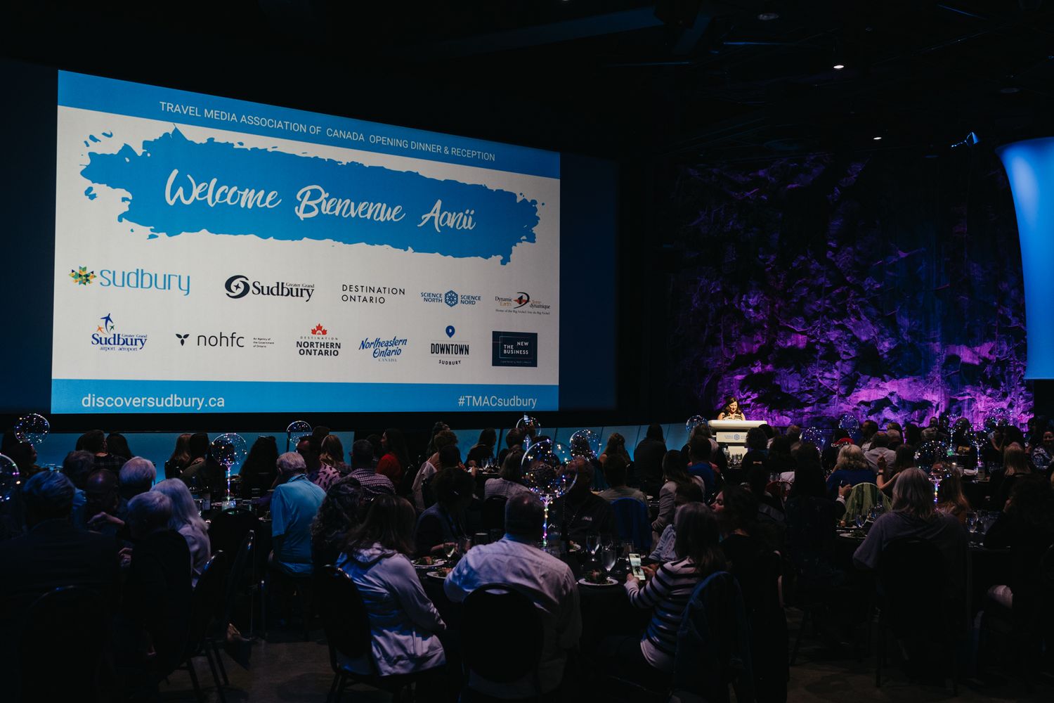 Corporate conference stage with blue lighting and sponsor logos displayed on screen.