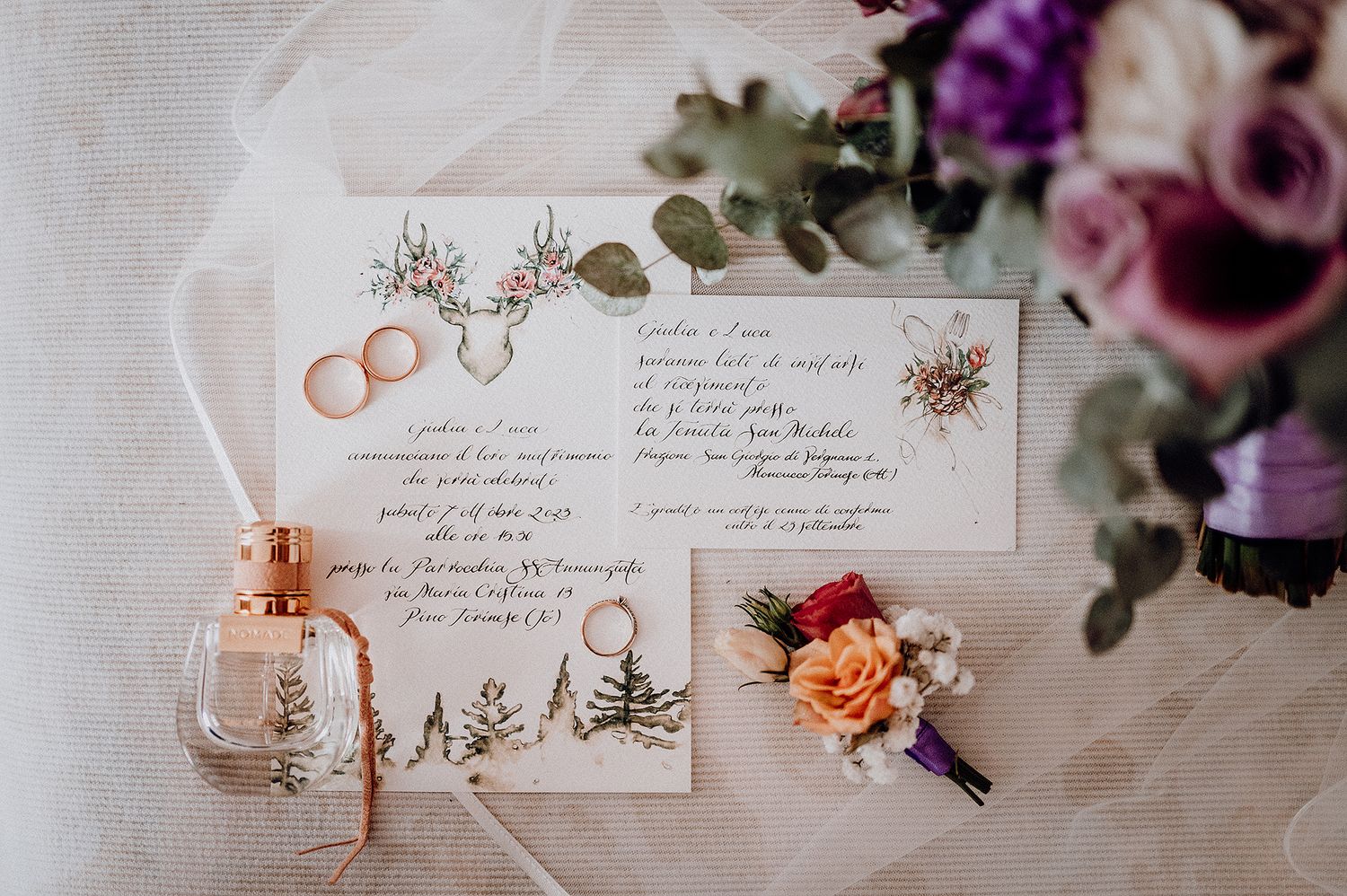 Wedding flat lay with floral invitation, gold rings, perfume bottle, and colorful boutonniere.