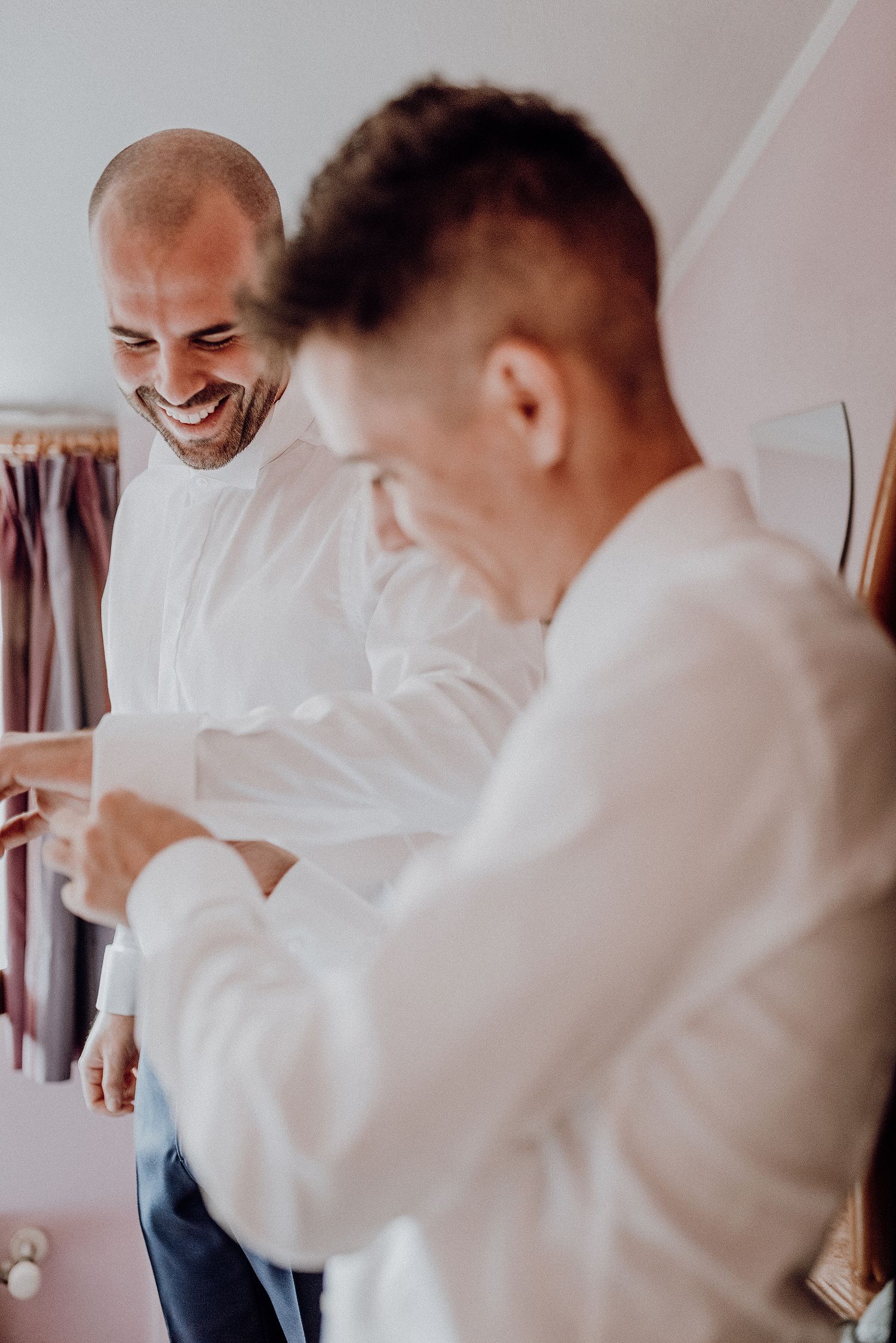 Two grooms smiling while helping each other dress in white shirts before wedding ceremony.