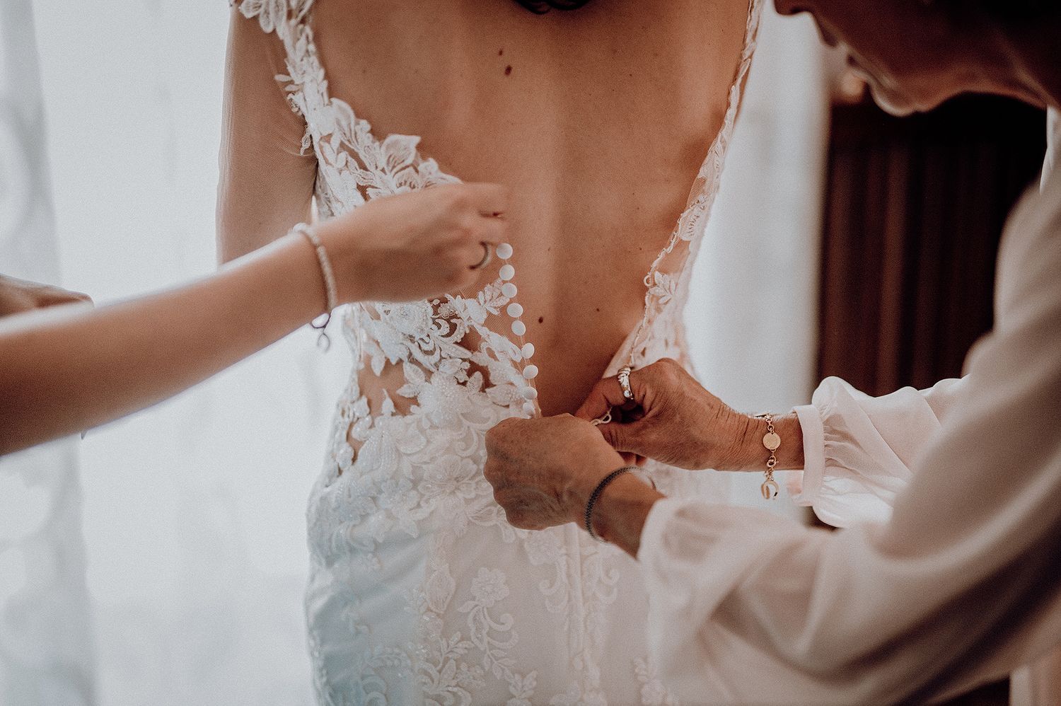 Wedding helper fastening buttons on back of lace bridal gown before ceremony.