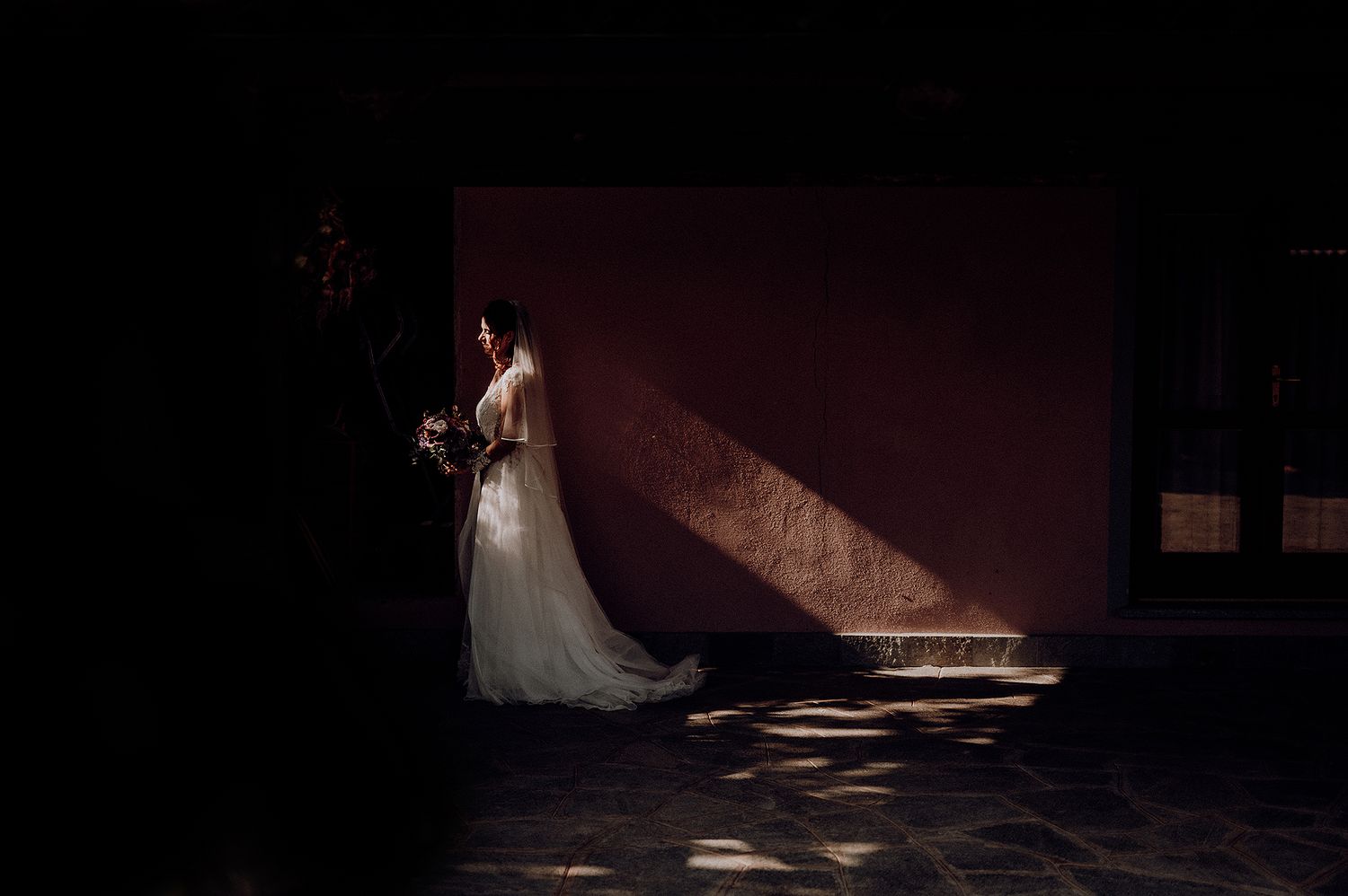 Bride in white gown holding bouquet stands in dramatic light against a terracotta wall.