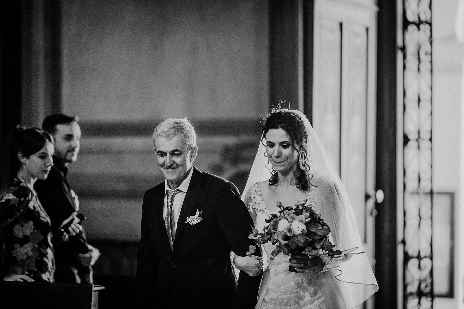 Bride walks down the aisle escorted by her father in a classic black and white wedding photo.