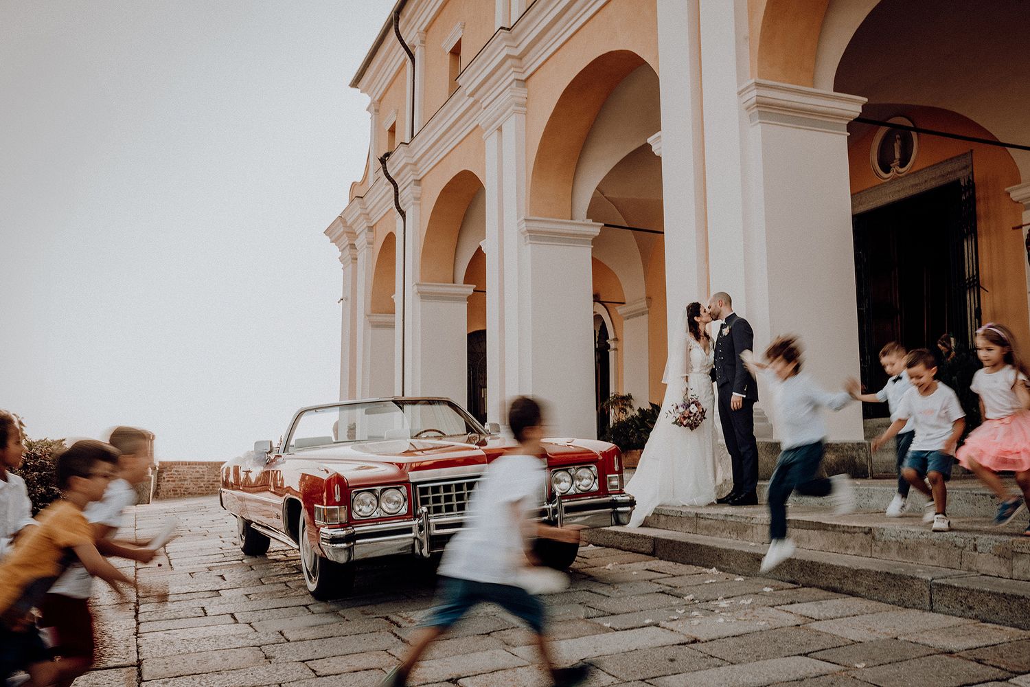 Newlyweds kissing beside a vintage red convertible outside a classic church with children playing.
