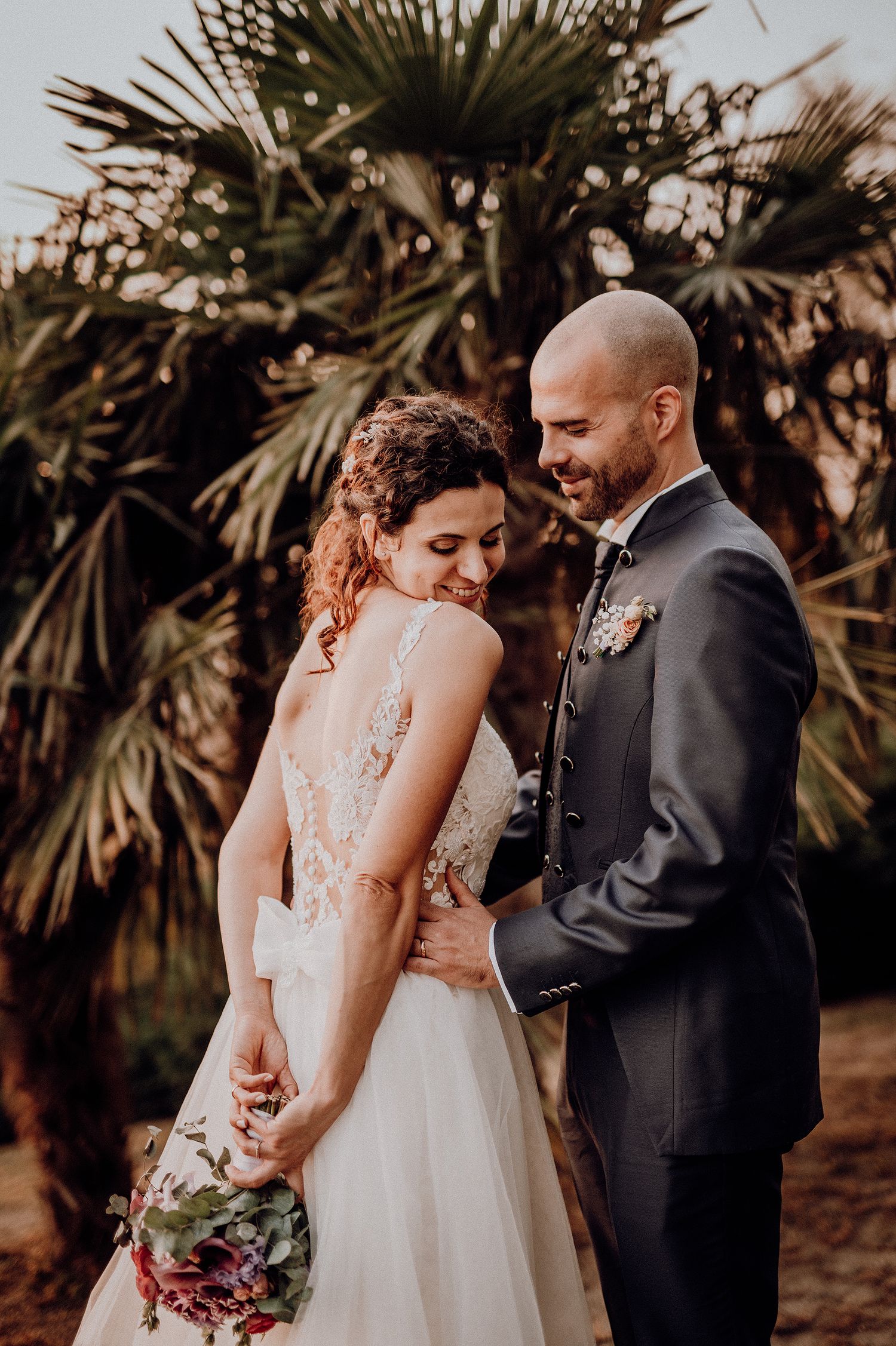 A smiling groom in a dark suit embraces his bride in a lace wedding dress outdoors.
