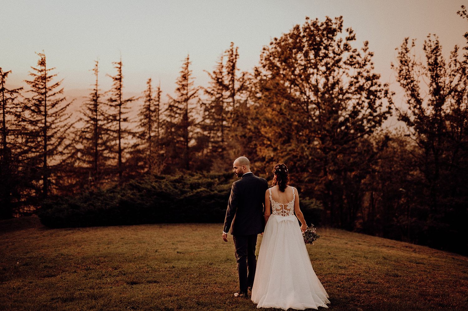 A bride and groom walk hand in hand through a scenic forest at golden hour sunset.