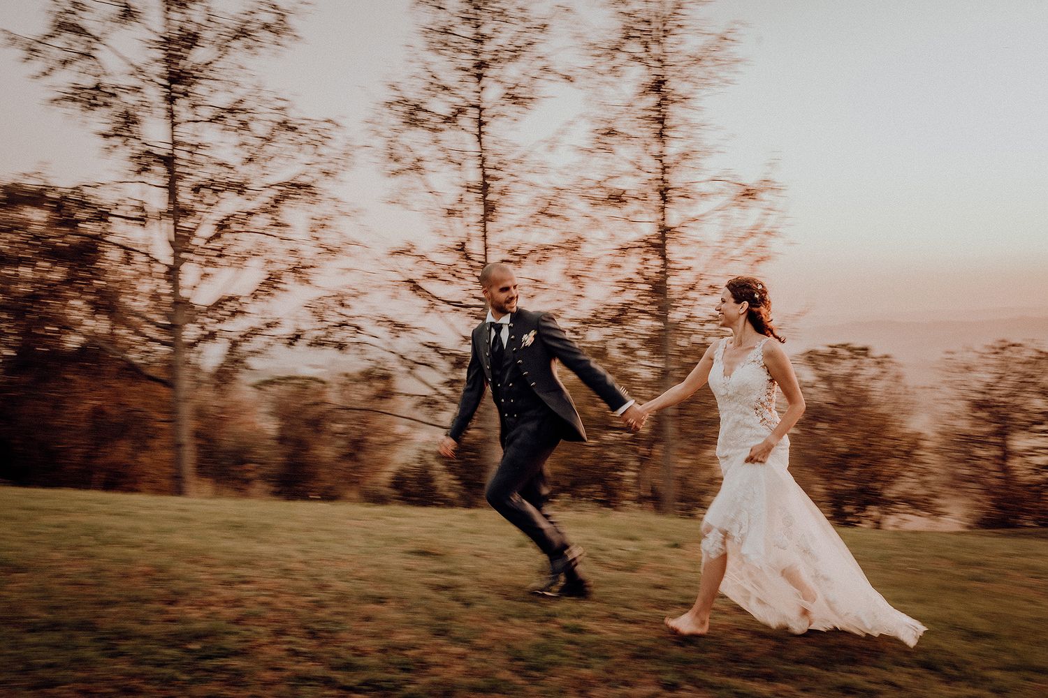 A joyful bride and groom run hand in hand across a green field at sunset.