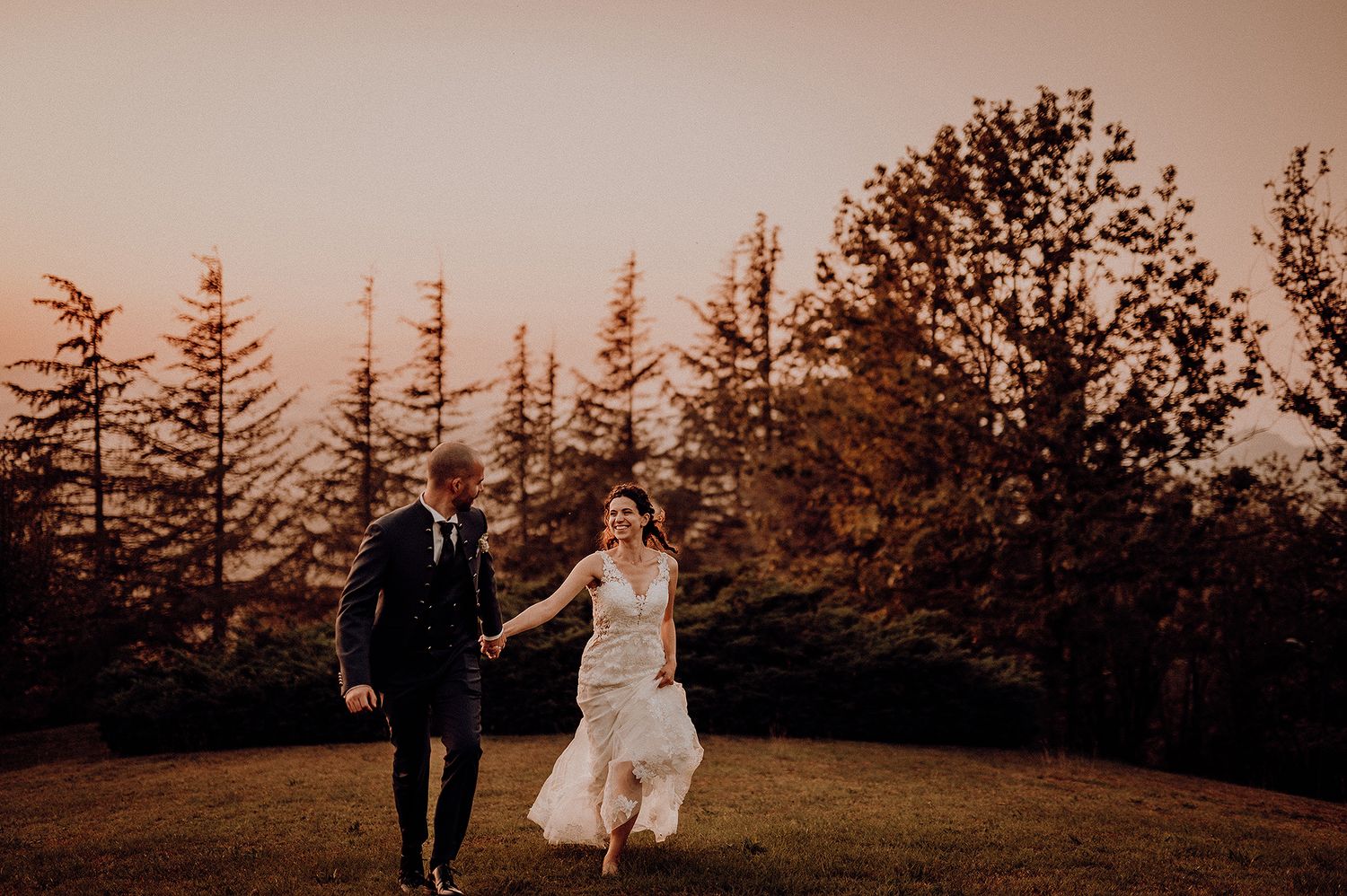A happy couple runs together through a sunlit outdoor wedding venue surrounded by trees.