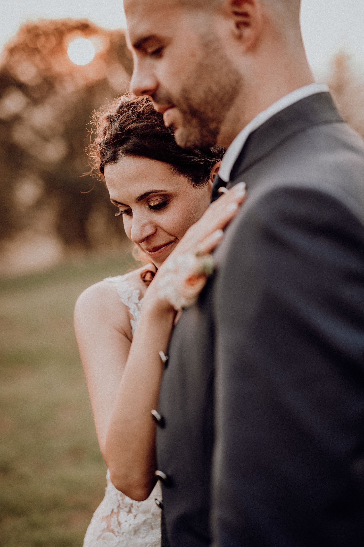 A bride in lace gown leans tenderly against her groom during a golden hour portrait.