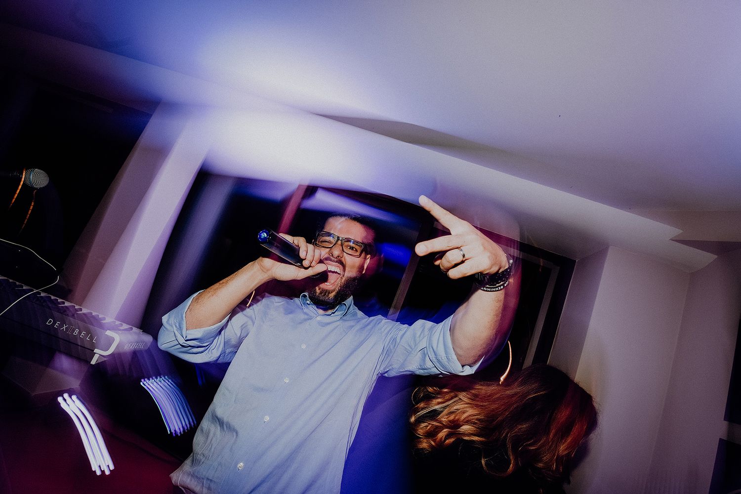 Man performing with microphone making hand gestures under purple stage lighting at party.