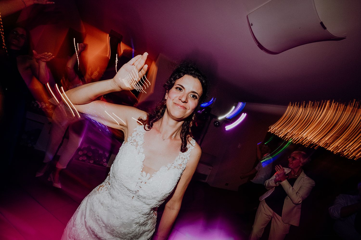 Bride in white lace dress dancing joyfully under colorful lights at wedding reception.