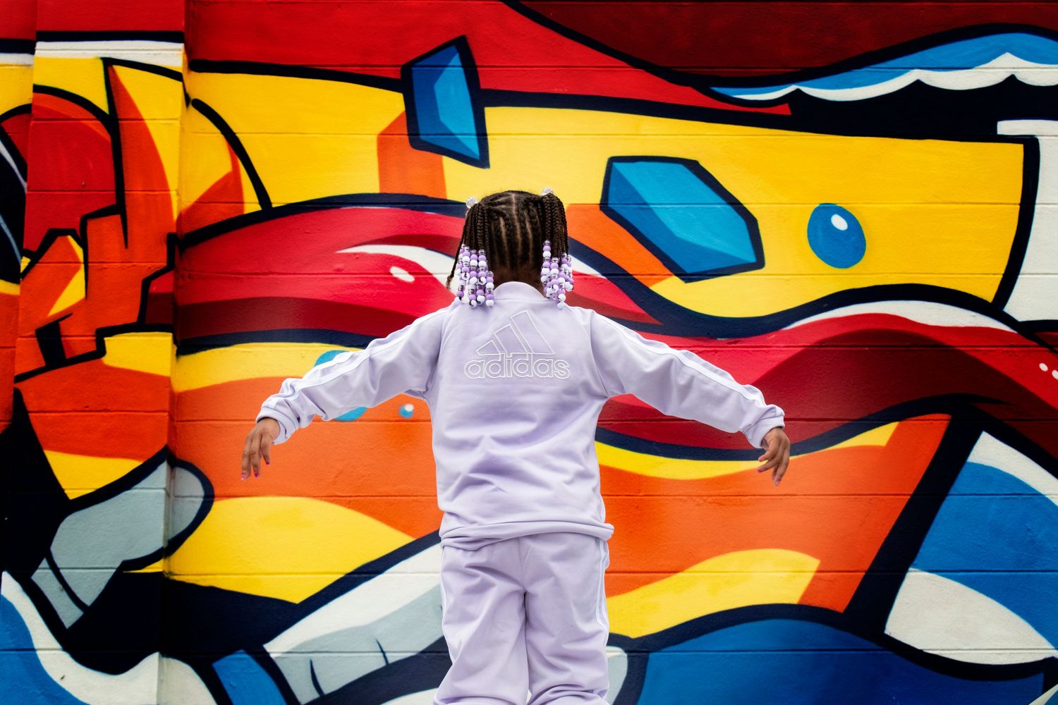 Child in white hoodie stands arms outstretched before a vibrant colorful graffiti mural wall.