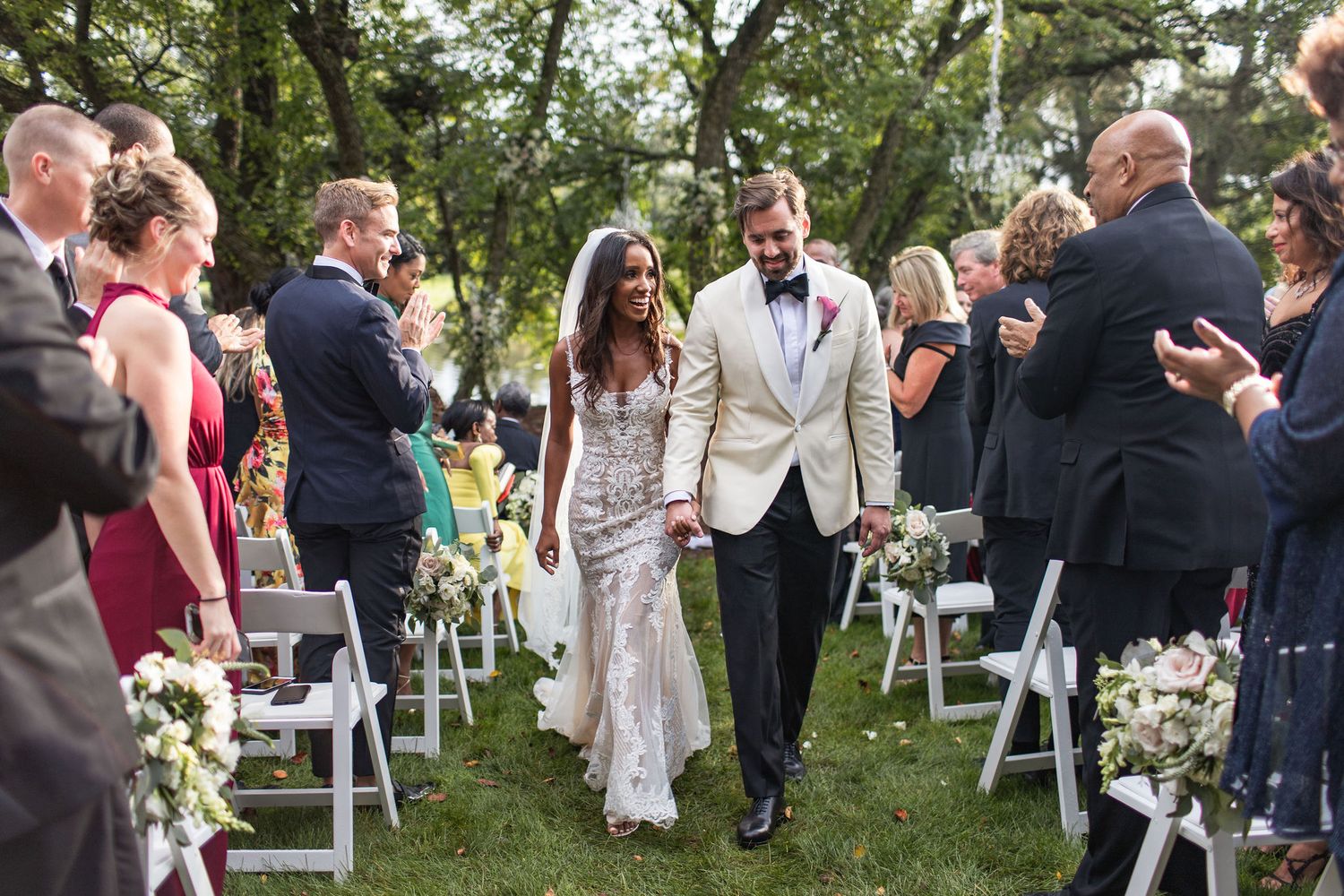 A newly married couple walks down the aisle at an outdoor garden wedding ceremony while guests celebrate.