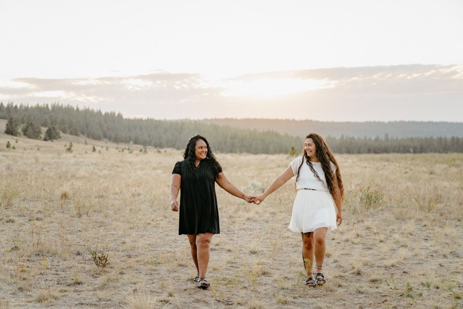 Two friends holding hands while walking through a scenic mountain meadow at sunset.