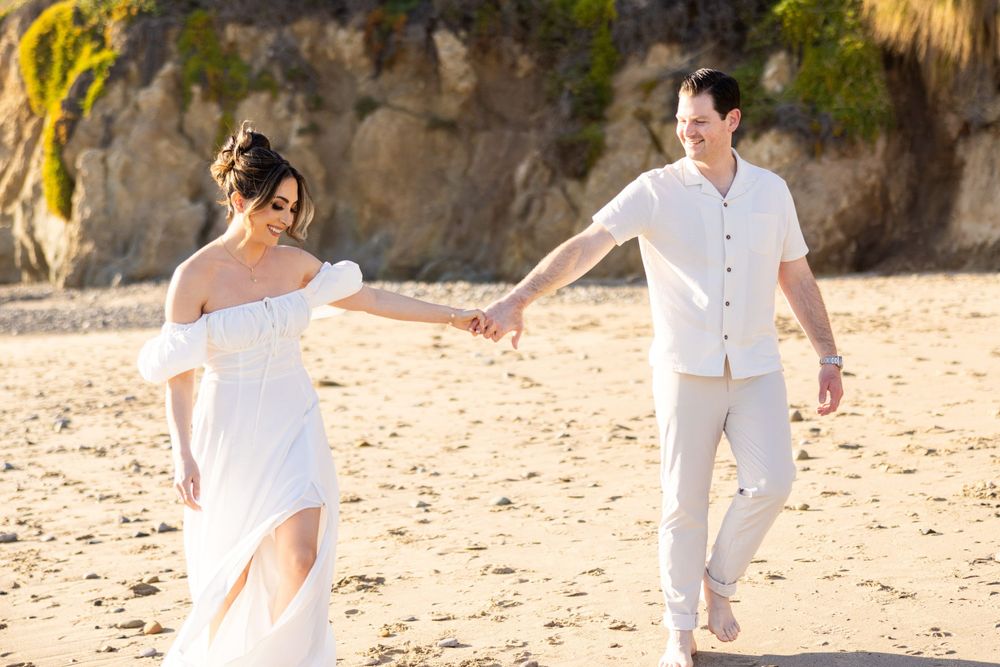 Couple in white attire walks hand in hand along sunny beach near cliffs.