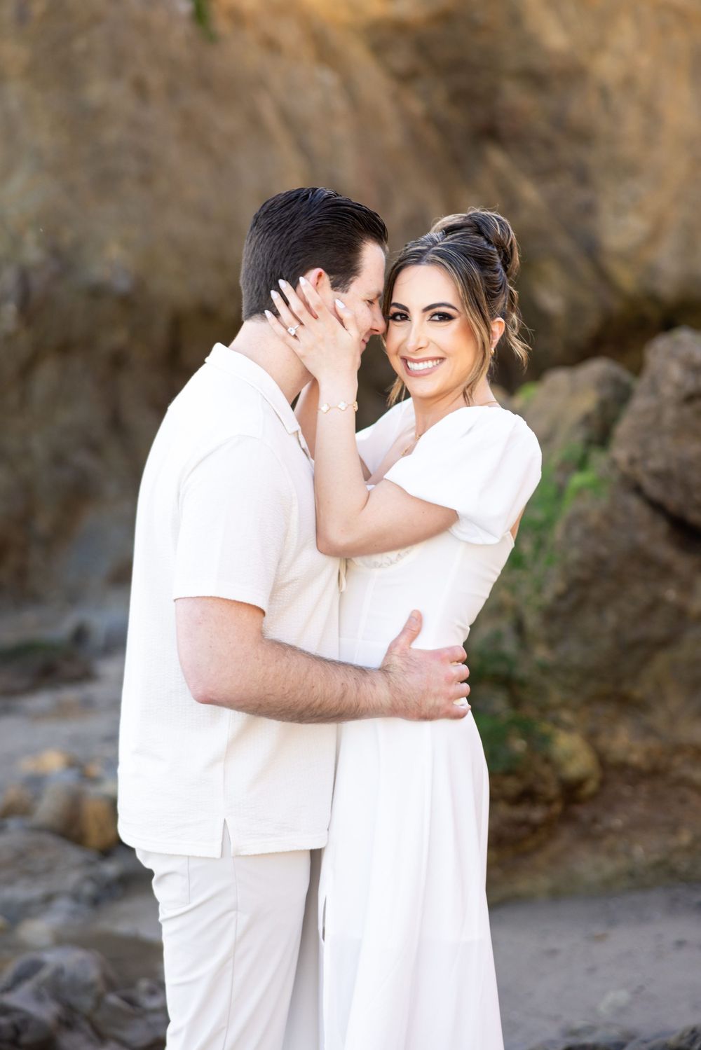 Intimate beach portrait of couple in white embracing against rocky coastal backdrop.