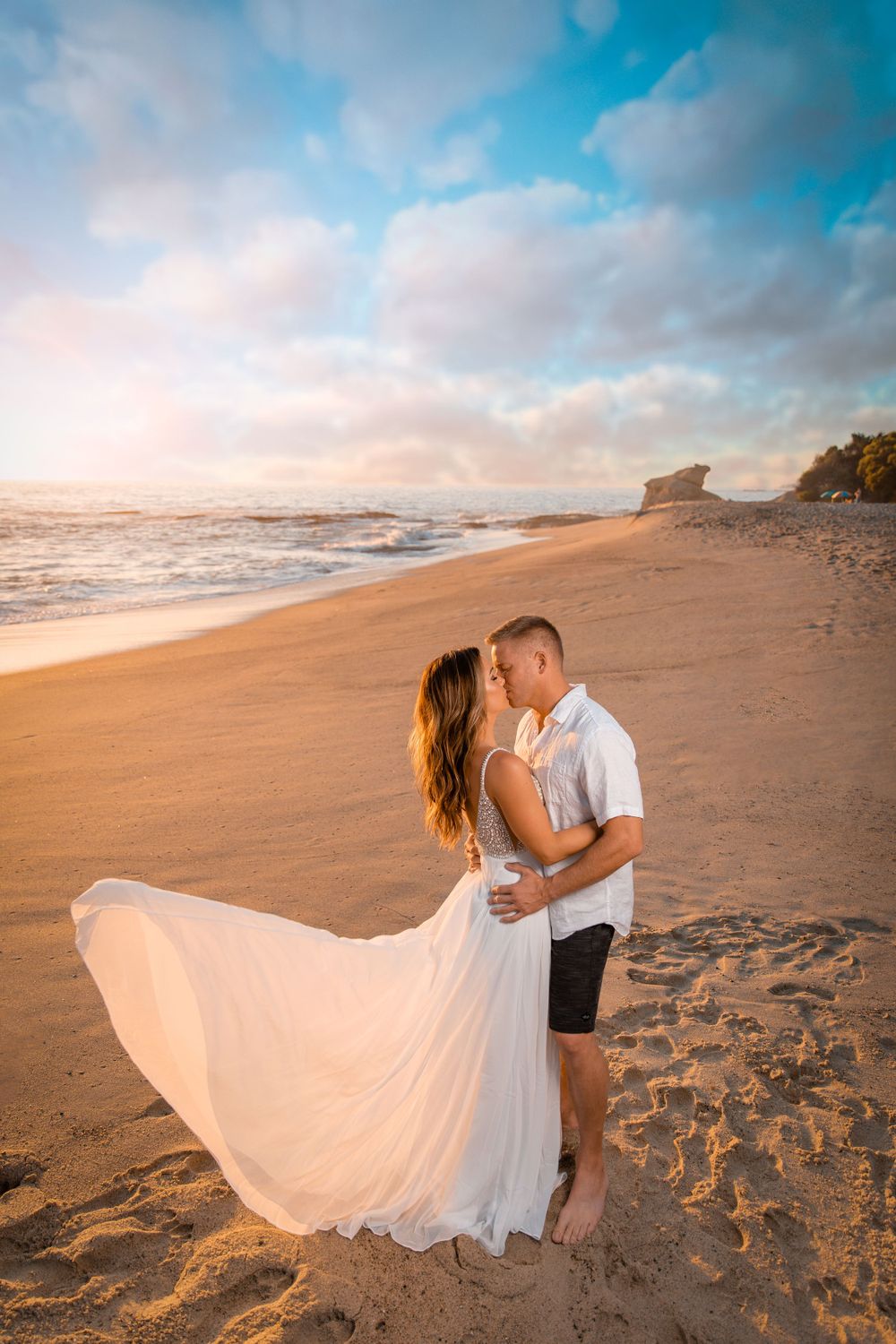 Couple in white outfits share intimate moment on sandy beach with flowing dress and sunset sky.