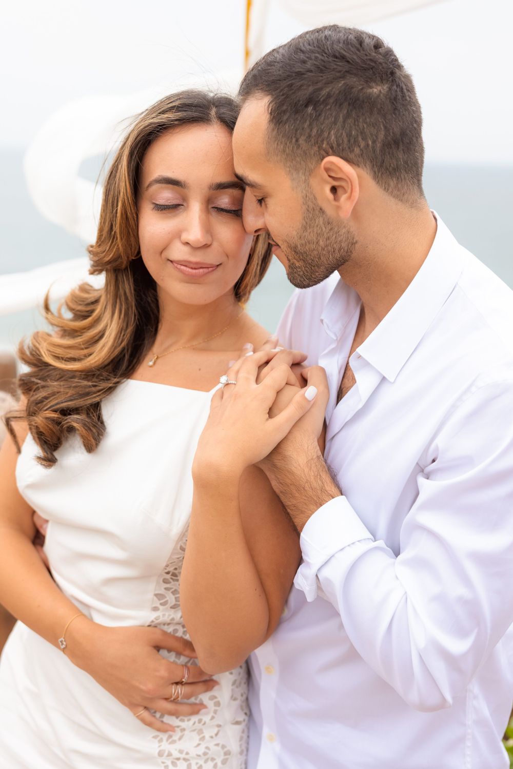 Intimate moment between couple in white outfits against scenic coastal background.