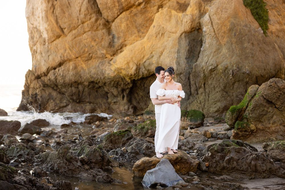 A couple in white attire stands on rocky coastal terrain against a dramatic cliff backdrop.