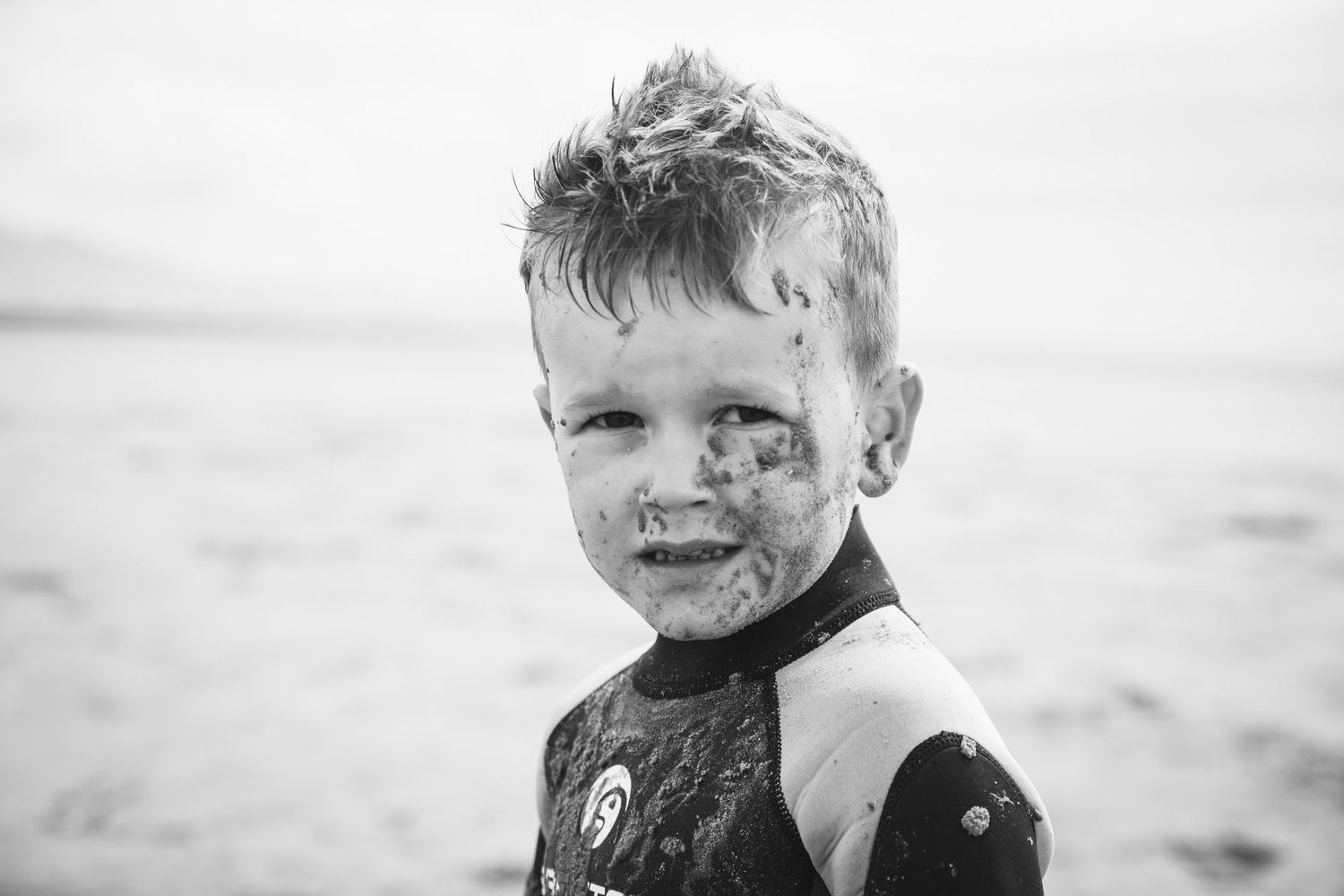 Black and white image of a child in a wetsuit with sand on their face at the beach.