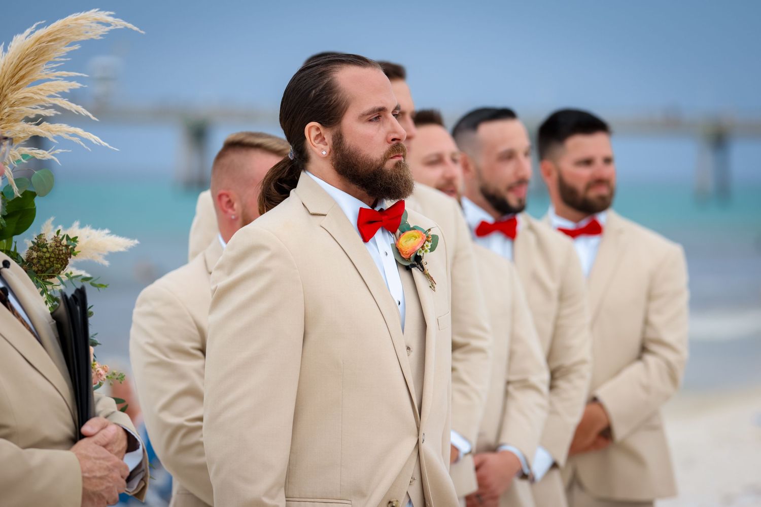 Groomsmen in beige suits and red bowties stand at a beachfront wedding ceremony.