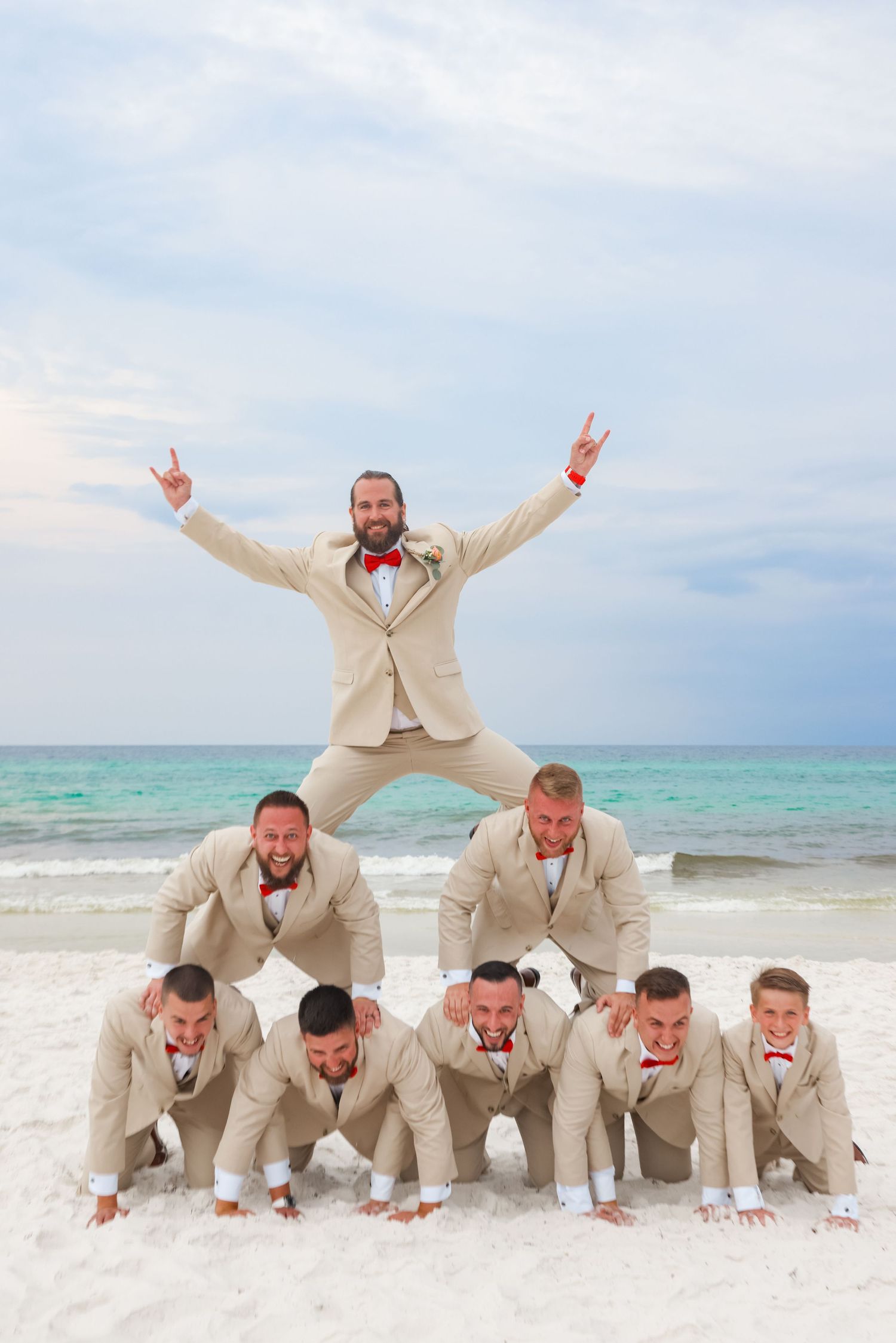 Fun groomsmen photo pyramid on beach with matching beige suits and red bowties.