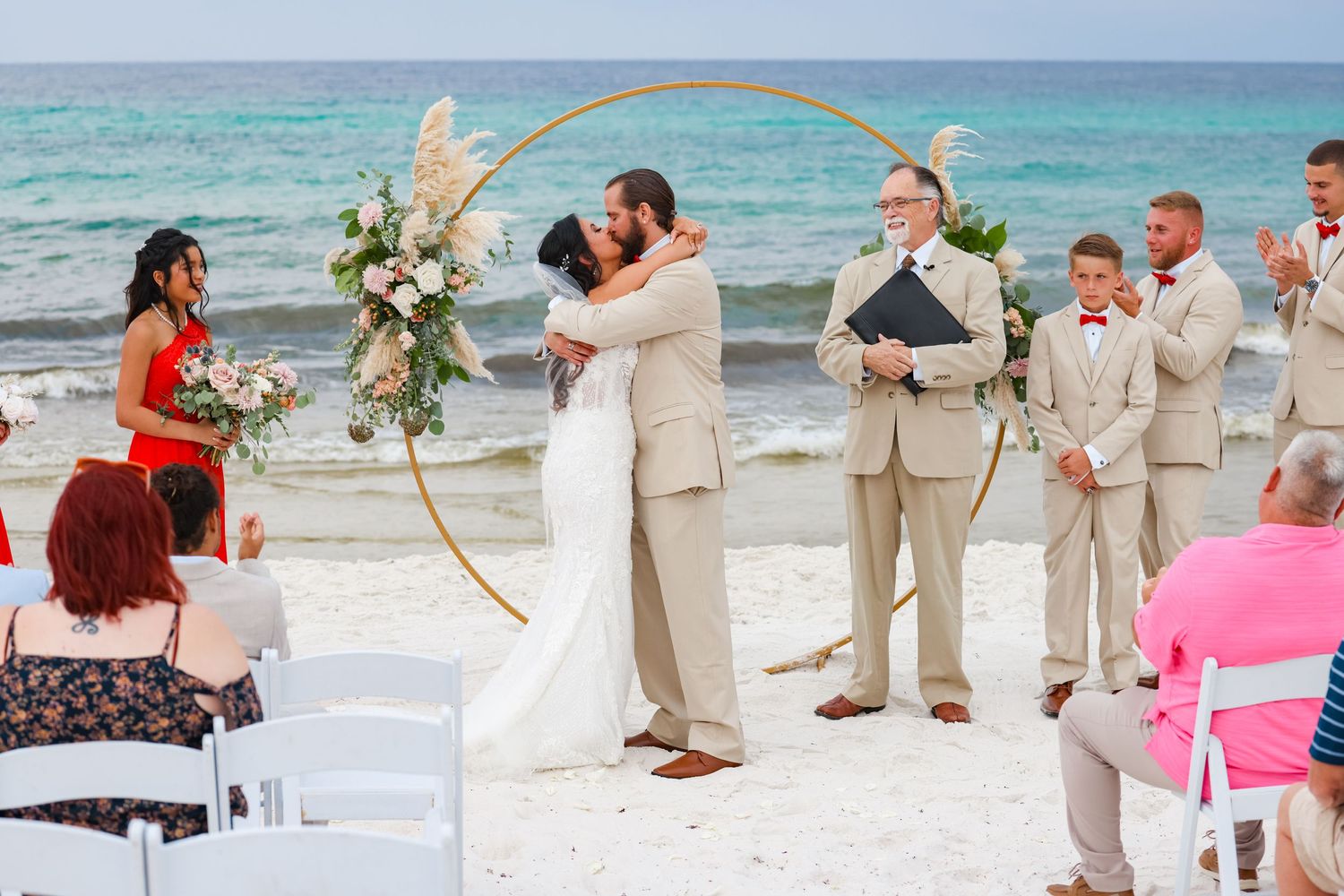 Beach wedding ceremony with circular arch, white chairs, and turquoise ocean backdrop.