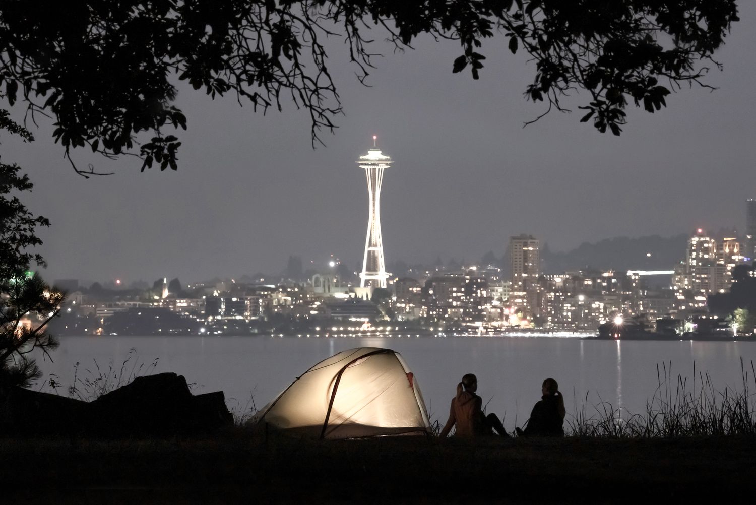 Illuminated Seattle Space Needle cityscape with glowing tent and campers silhouetted against the water at night.