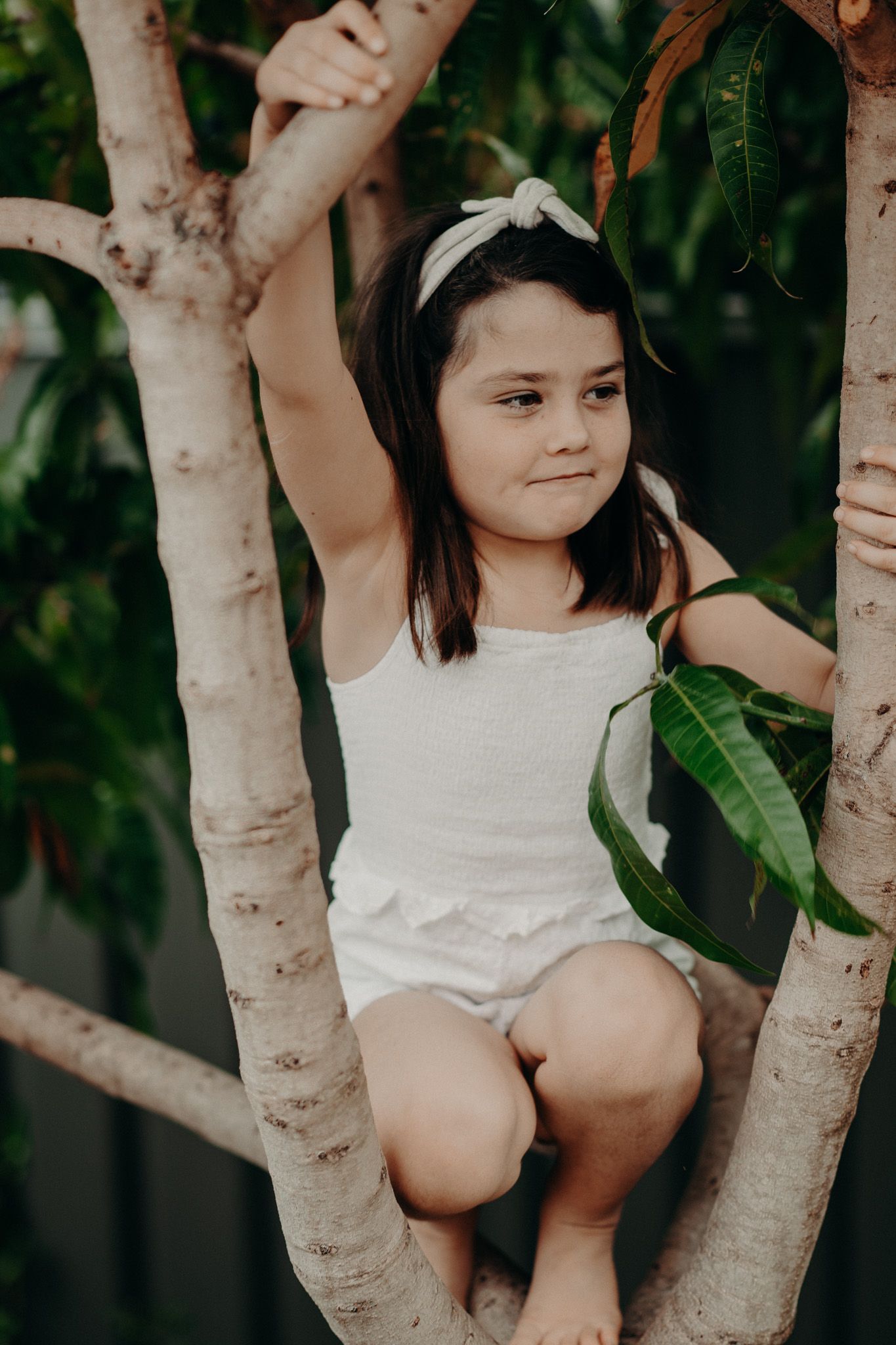 Isla climbing a tree wearing a white jumpsuit and headband.
