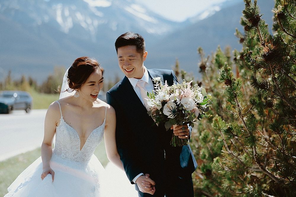 A wedding couple walks together in front of stunning mountain peaks with the bride carrying a white floral bouquet.