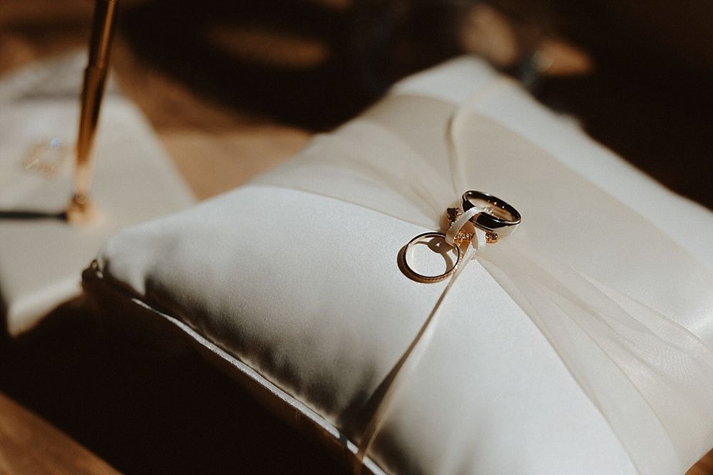 Wedding rings rest on a white silk pillow in dramatic lighting.