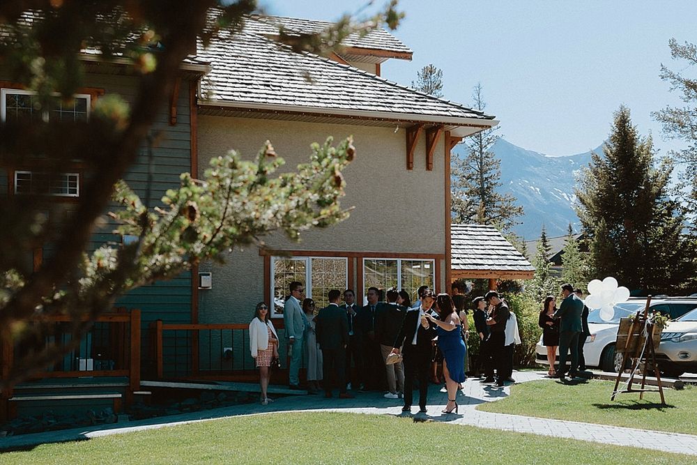 Wedding guests mingle outside a rustic mountain lodge with evergreen trees in the background.