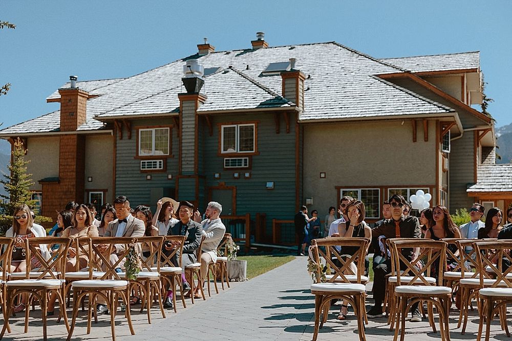 Outdoor wedding ceremony with wooden cross-back chairs and mountain lodge backdrop.