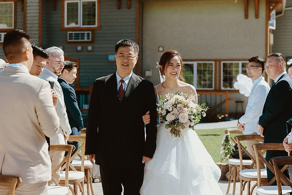 Outdoor wedding processional with wooden chairs and guests along the aisle.