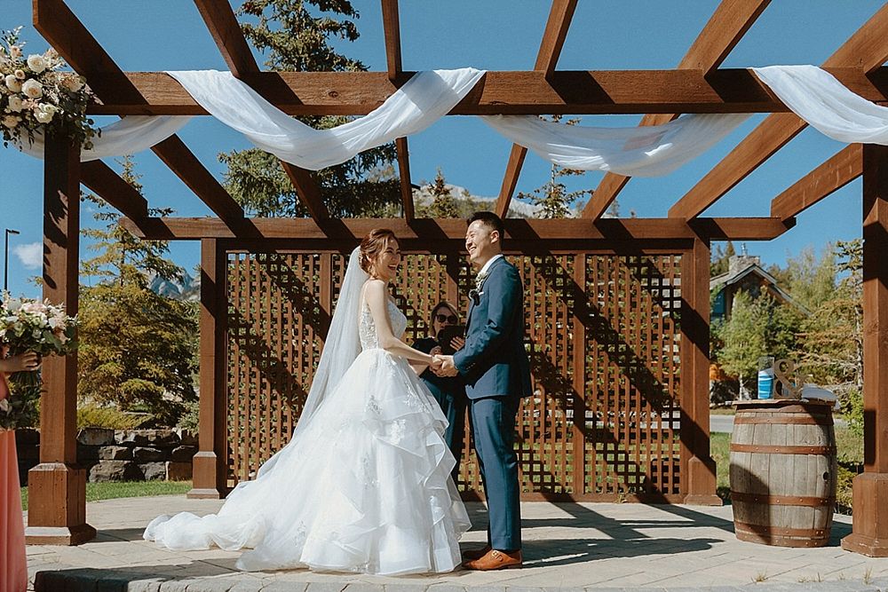 Wedding ceremony under a wooden pergola decorated with white fabric draping.