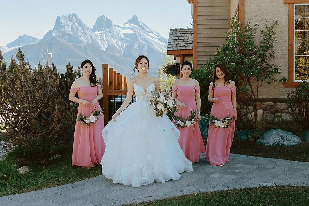 Bride in white gown with bridesmaids in pink dresses against mountain backdrop.