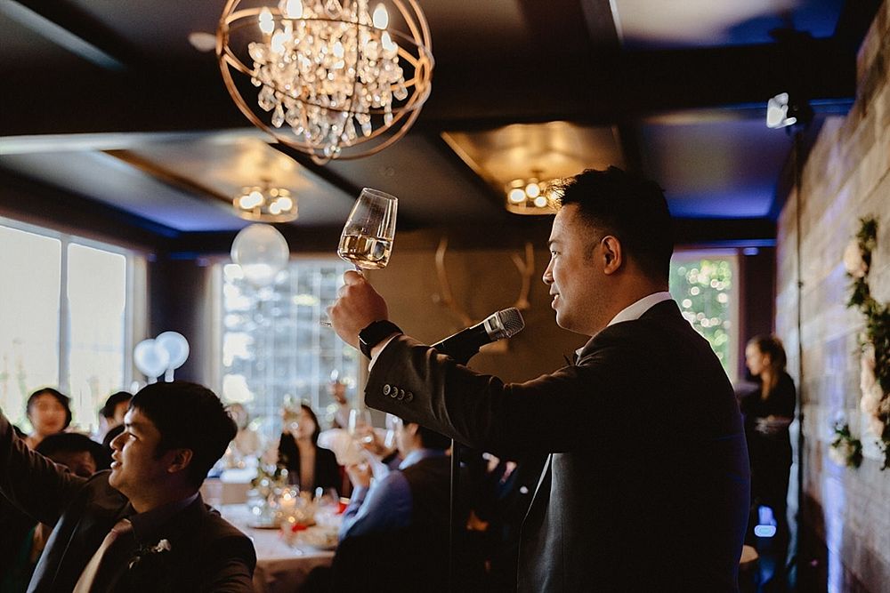 Crystal chandeliers cast warm light over wedding reception guests in rustic dining space.
