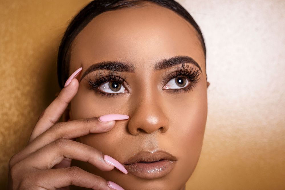 Close-up of a Black woman with long lashes, arched brows, and pink almond nails framing her face.