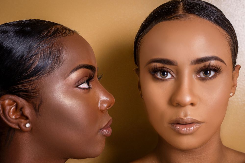 Two Black women with flawless makeup pose together against a gold background in a beauty portrait.