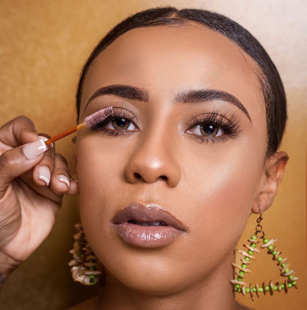 Black woman applying eye makeup with a brush, wearing green earrings and glossy lips.