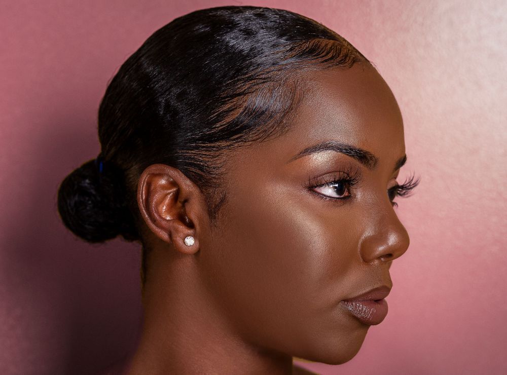 Side profile of a Black woman with a bun, pearl earring, and flawless skin on a pink background.