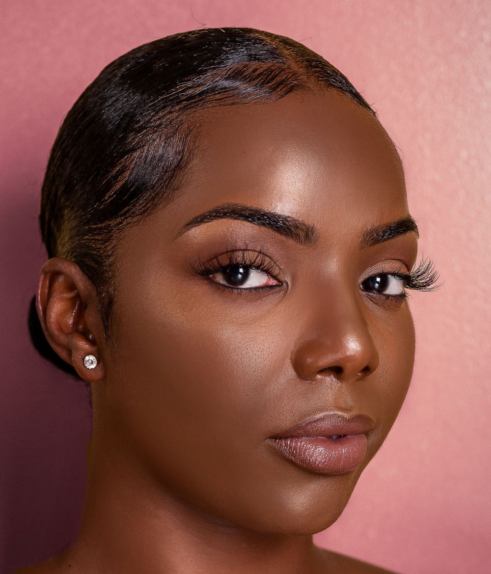 Black woman with sleek hair, diamond stud earring, and natural glam makeup against a pink background.