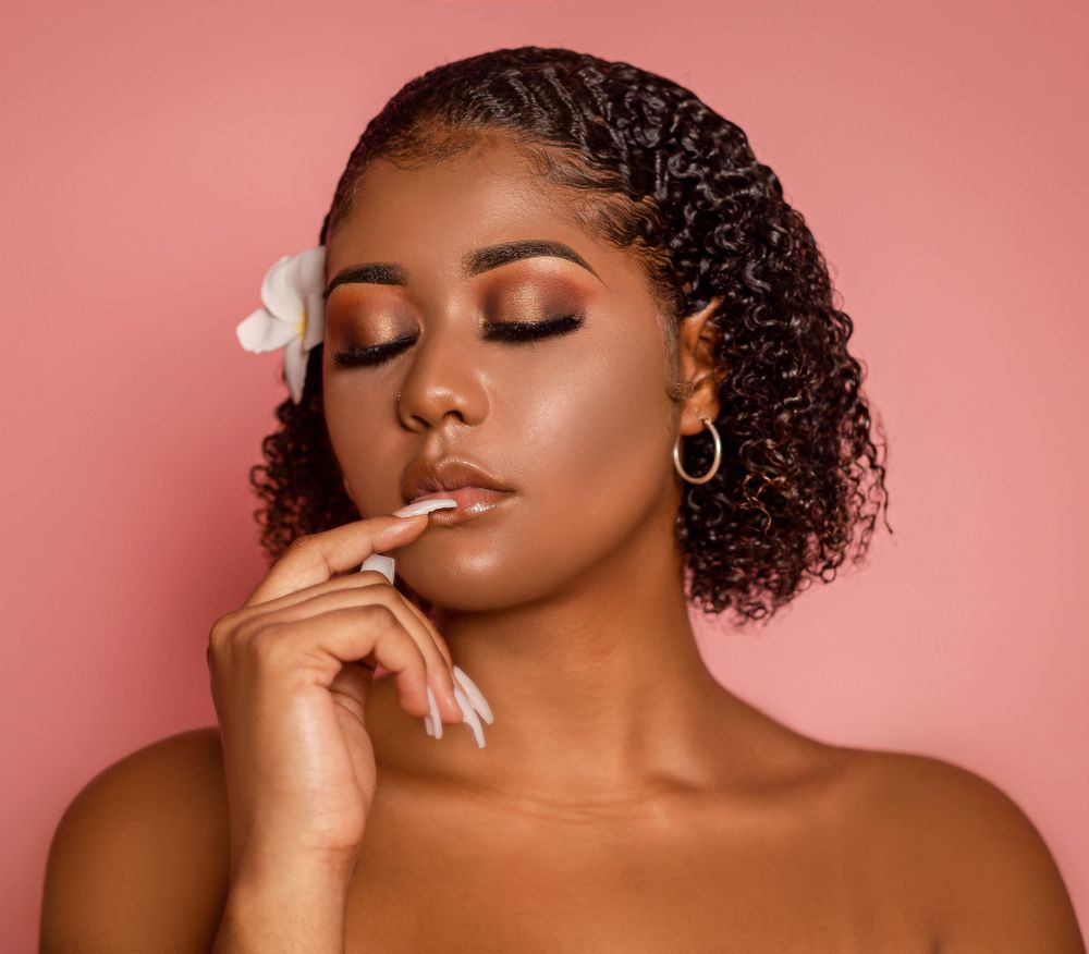Black woman with curly hair and a white flower, wearing warm eye makeup, eyes closed on pink backdrop.