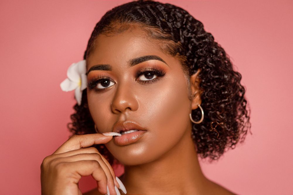 Black woman with curly hair, white flower accessory, and bold eye makeup posing on a pink background.
