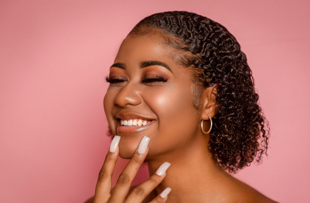 Smiling Black woman with curly hair, hoop earring, and natural nails against a soft pink background.